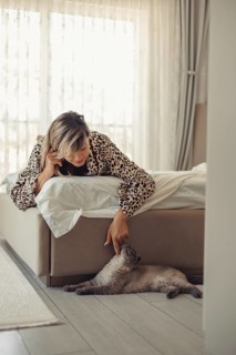 A woman, wearing a leopard print robe, leans over the edge of a bed to lovingly play with her cat