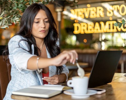 A woman in a cozy cafe stirs a cup of coffee while working on her laptop. She is surrounded by a