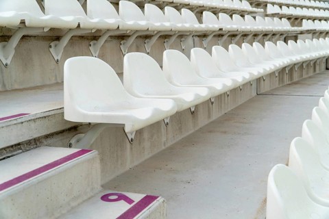 Rows of empty white seats await spectators in an outdoor athletics stadium, with visible seat