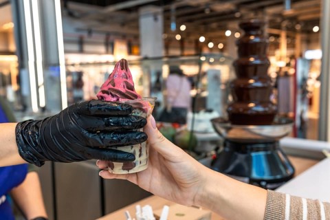 Vendor's gloved hand serving a pink and chocolate swirl soft serve ice cream in a cup to a