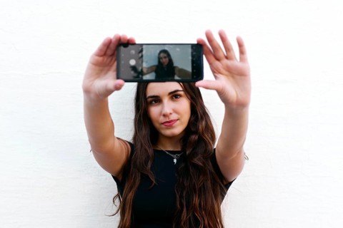 A young woman with long hair takes a selfie with her smartphone against a plain white wall. She
