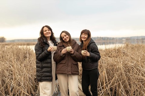 Three female friends smile joyfully as they hold mugs, standing together in a tranquil outdoor