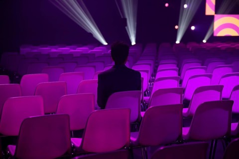 A indian man sits in an empty auditorium filled at a conference of AI in Amsterdam with purple lit
