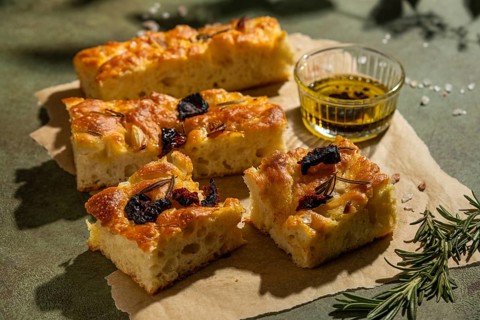 Golden homemade focaccia slices topped with olives and rosemary, accompanied by a small dish of