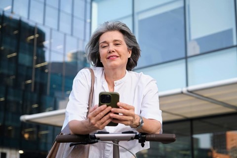 Confident mature woman in a white blouse stands outdoors, smiling while using her smartphone on an