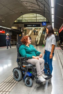 Vertical photo of a disabled woman in wheelchair talking to an asian female friend waiting at metro