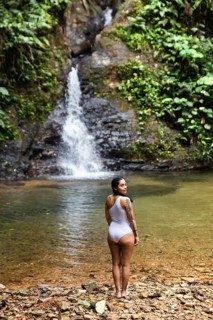 Back view of a young Ecuadorian woman in a white swimsuit stands admiring a striking waterfall in