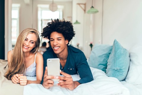 A young couple relaxes on a bed, smiling as they browse a smartphone. The cozy bedroom setting with