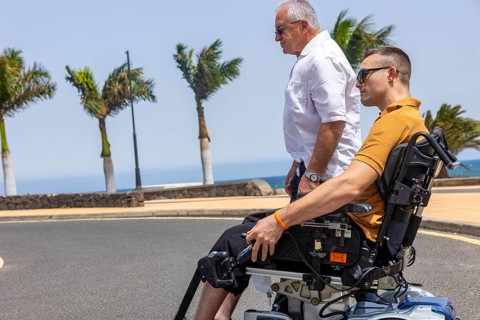 A young man in a wheelchair accompanied by an older man walking beside him, both enjoying a sunny