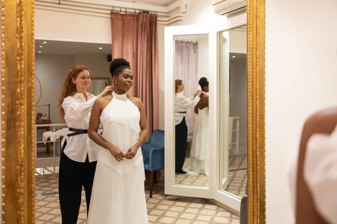 A bride stands in front of a mirror in a fitting room while a stylist adjusts her white wedding