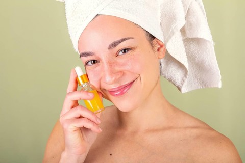 A woman with a towel wrapped around her head holds a small bottle of facial serum. Her radiant