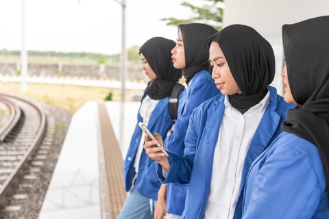 A group of Asian Muslim women in blue uniforms wait at a train station, symbolizing their return to