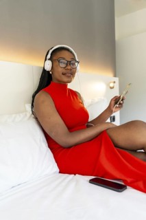 Businesswoman wearing red dress is relaxing on a hotel bed, listening to music with headphones and