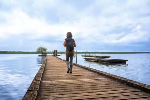 Woman walking with backpack on a wooden pier extending into lake azur under a cloudy sky,