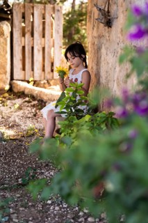 A young girl in a white dress admires a sunflower while sitting in a rustic outdoor garden. The