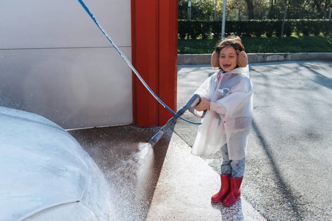 A joyful child in a raincoat and earmuffs uses a hose to spray water on a car, beaming with delight