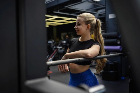 A fit blonde woman rests on gym equipment, wearing blue leggings and a black top. The gym setting