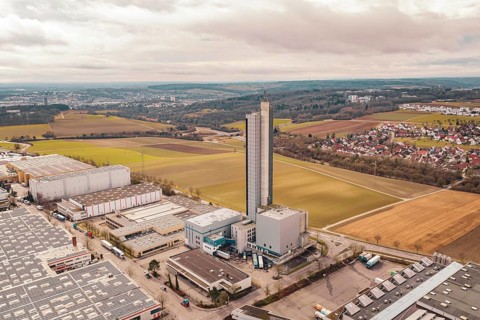 Tall industrial building surrounded by fields and a village in the background under cloudy sky,