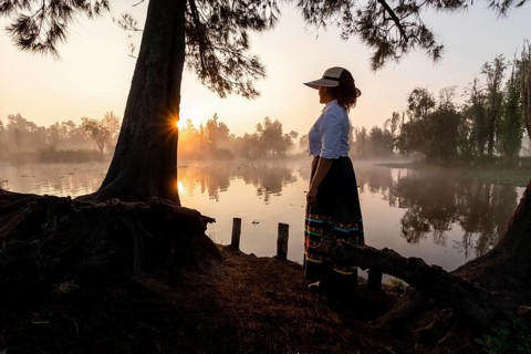 A woman in traditional attire stands by the water in Xochimilco, Mexico, as the sun rises,