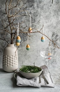 An Easter table setting featuring a ceramic vase with suspended painted eggs from bare tree