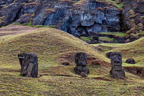 Three stone Moai statues in front of a rock formation in grassy surroundings, The Moai statues on