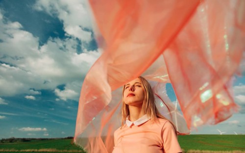 A woman stands in a field with her eyes closed, enveloped by a flowing pink fabric. The sky is