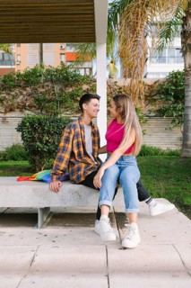 A lesbian couple shares a joyful moment on a bench outdoors. Behind them, a bag with the Pride flag