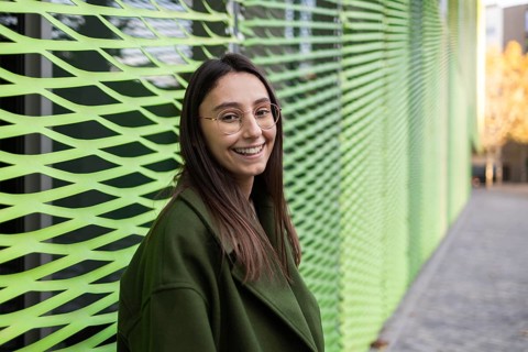 A woman, wearing glasses and a green coat, smiles warmly while leaning against a modern green wall