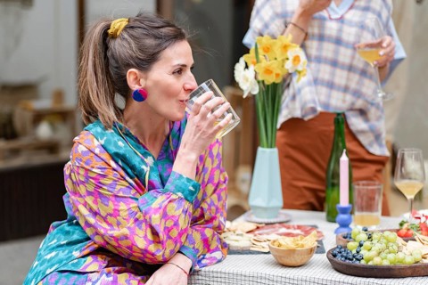 Woman in colorful attire enjoying a drink at an outdoor brunch. A table with fresh fruits, snacks,