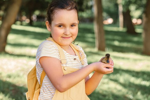 A smiling little girl tenderly holds a cute duckling in her hands outdoors looking at camera. The