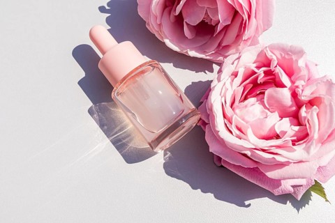 Top view of a glass dropper bottle filled with rose water serum, placed alongside two pink roses on