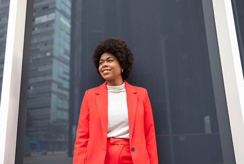 A woman in a bright red suit stands confidently against a modern urban backdrop. Her smile and