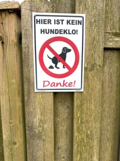 Sign with pictogram and inscription Hier ist kein Hundeklo on the outside of a wooden fence,