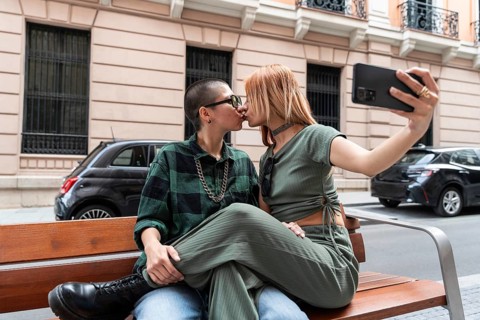 A Colombian lesbian couple captures a romantic moment with a selfie while kissing on a city bench.