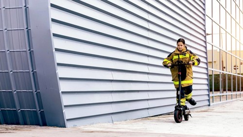 A girl wearing a firefighter suit rides an electric scooter against an urban backdrop with a modern