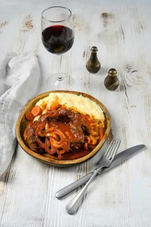 A wooden bowl with a red-brown beef stew, garnished with sliced onions and carrots, sits next to a