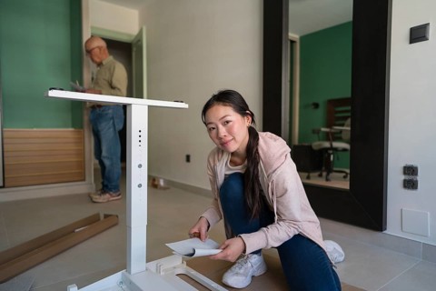 A young Asian woman kneels on the floor, carefully reading the manual while assembling a white desk