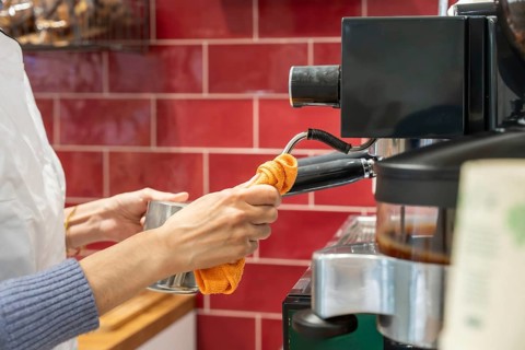Barista wiping steam wand of espresso machine with orange cloth, preparing fresh milk froth, close