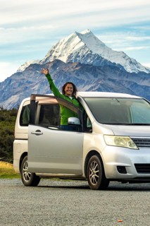 A woman stands joyfully by her car in autumn at Mount Cook, New Zealand. The majestic mountain