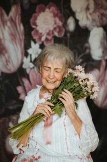 An elderly woman with a joyful expression holds a bouquet of flowers, surrounded by a floral