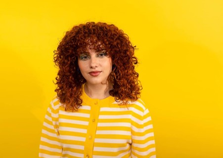 Portrait of a woman with vibrant, curly red hair wearing a yellow and white striped sweater, set