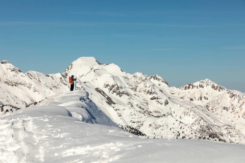 A snowboarder with a bright orange backpack stands atop a snowy mountain ridge, overlooking a vast