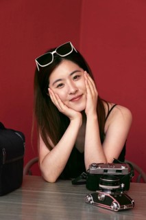 A young asian woman sits at a table with a vintage camera, sporting sunglasses on her head. The red