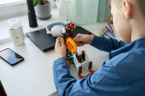 A young child focuses intently on assembling a colorful robotic toy on a desk. Surrounding him are