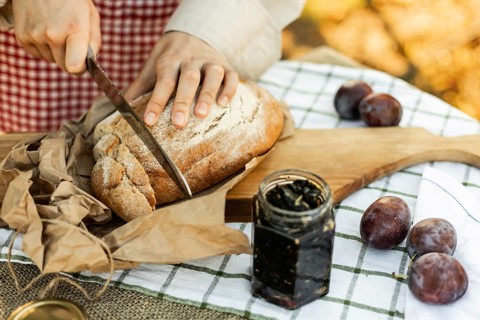 Hands slicing rustic bread on a wooden board, surrounded by fresh plums and jam. An inviting scene