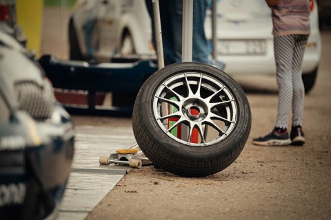 A close-up of a racing tire resting on the ground in a motorsport pit stop area. The scene includes