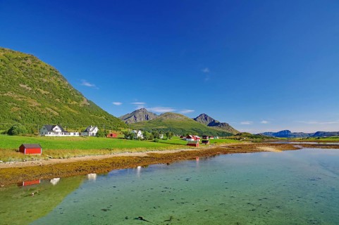 Clear water reflects the small village and surrounding mountains under clear skies, Leknes,