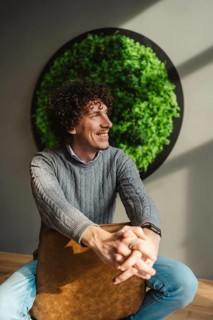 A young man with curly hair smiles while sitting against a lush green wall. The image emphasizes