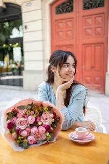 A woman sits at an outdoor cafe table, smiling with delight as she admires a beautiful bouquet of