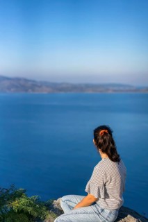 A woman in casual attire sits on a rocky outcrop, gazing at the tranquil blue sea under a clear sky